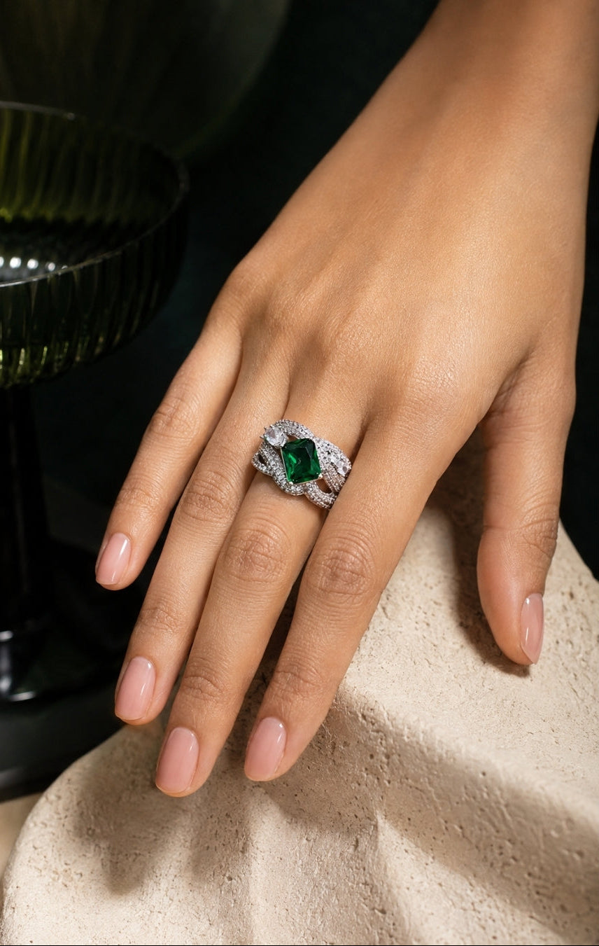 A close-up of a hand wearing a silver ring with a marquise-cut green gemstone and small clear accent stones, resting on a textured beige surface.