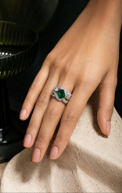 A close-up of a hand wearing a silver ring with a marquise-cut green gemstone and small clear accent stones, resting on a textured beige surface.