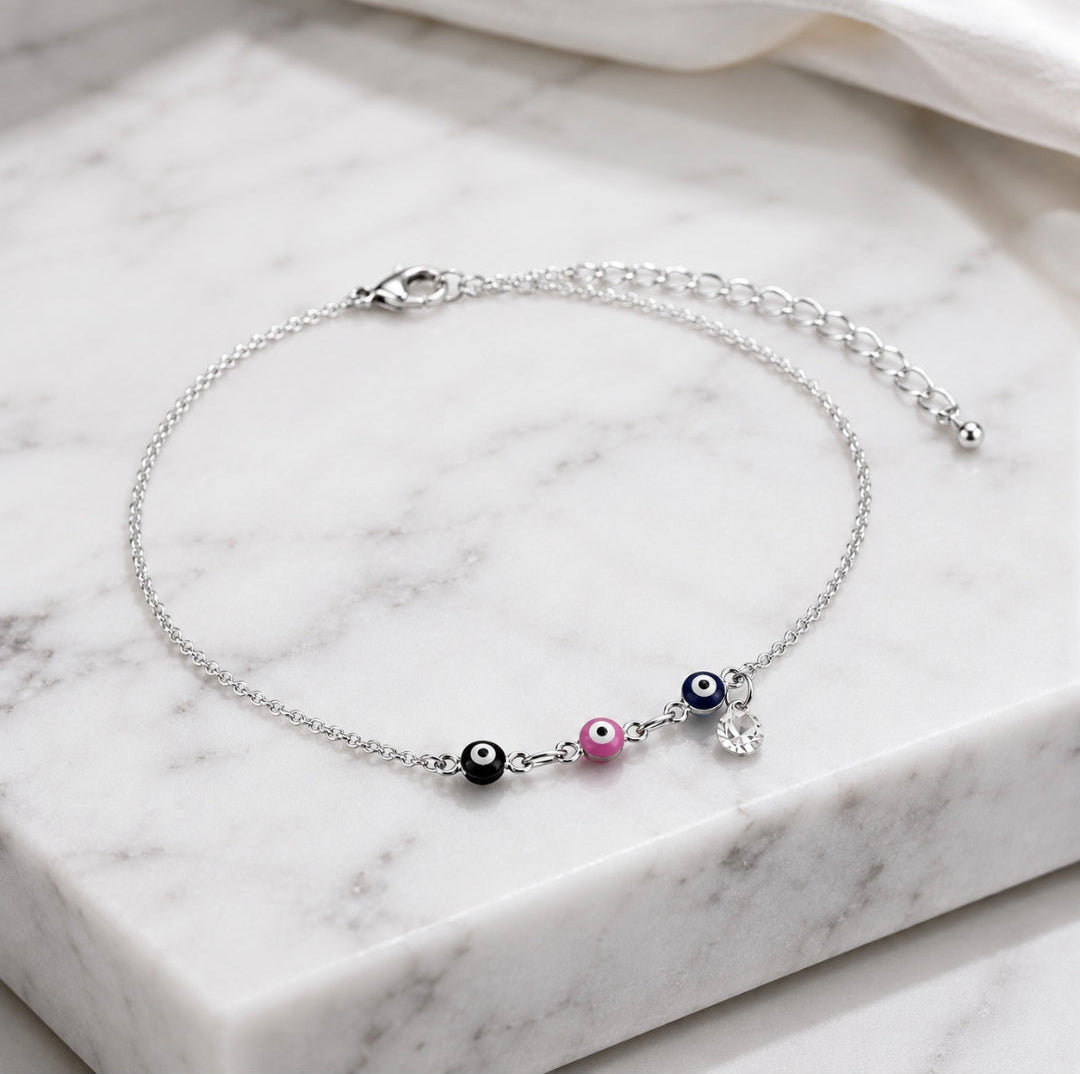 A delicate silver chain anklet with three evil eye beads (black, white, pink) and a small silver heart charm, displayed on a white marble block.