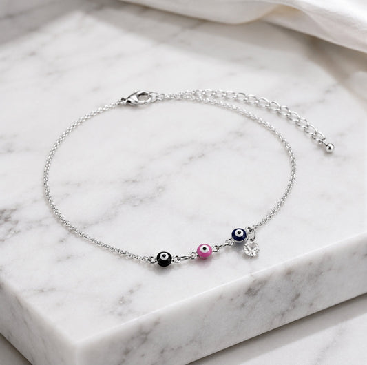 A delicate silver chain anklet with three evil eye beads (black, white, pink) and a small silver heart charm, displayed on a white marble block.