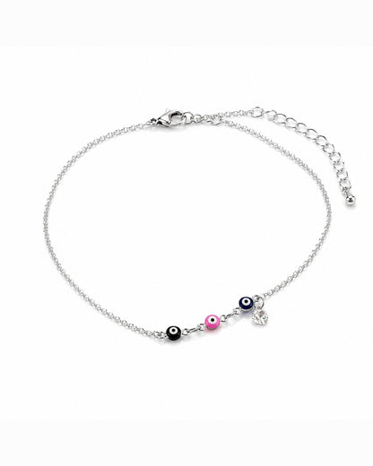 A delicate silver chain anklet featuring three evil eye charms in black, pink, and blue, with a small silver heart charm, displayed on a clean white background.