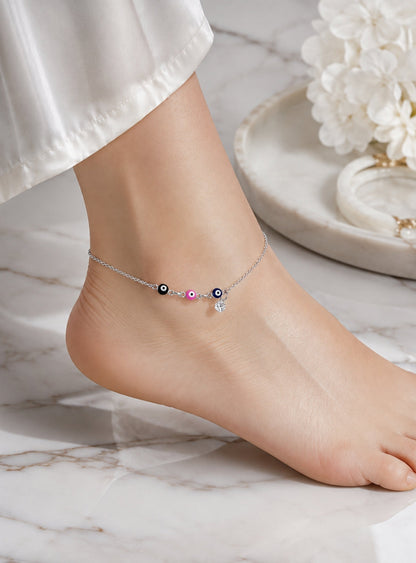 A delicate silver anklet with evil eye beads and a small dangling charm, shown worn on a person's ankle. The foot is resting on a white marble surface with white flowers in the background.
