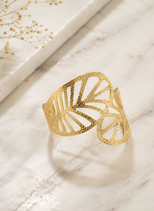A textured gold cuff bracelet with an openwork leaf pattern, displayed on a light marble surface with dried baby's breath flowers in the top-left corner.