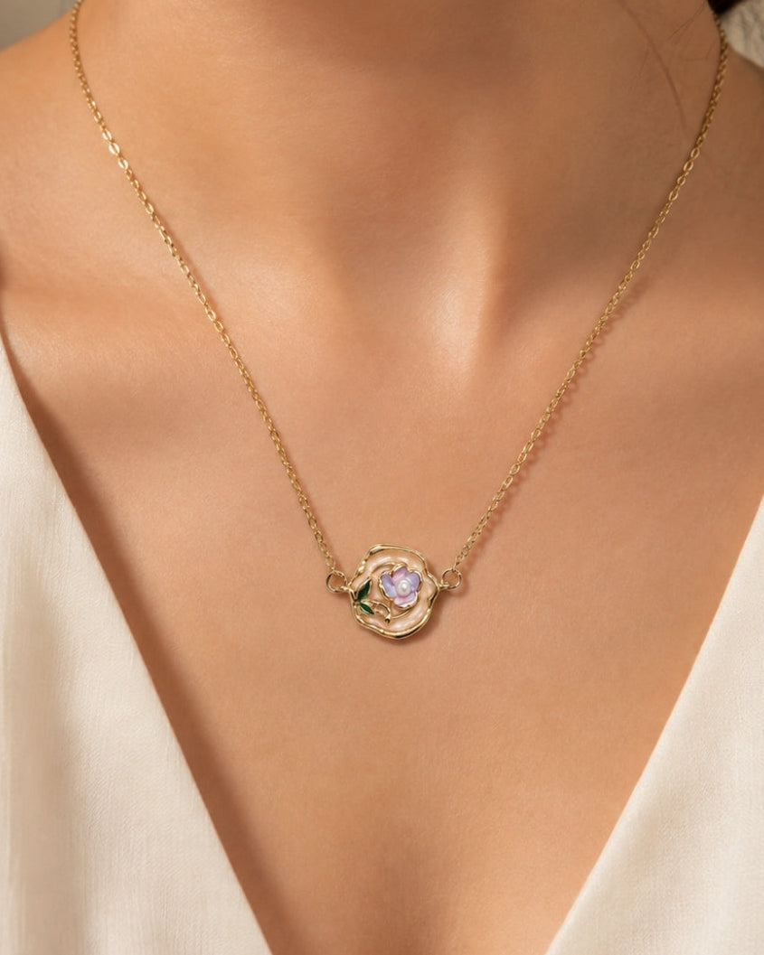 A close-up of a model's neck and chest wearing a delicate gold chain necklace with a small flower-shaped pendant featuring a central iridescent stone and green accents.