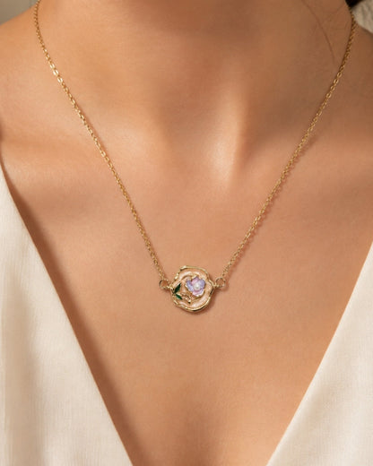 A close-up of a model's neck and chest wearing a delicate gold chain necklace with a small flower-shaped pendant featuring a central iridescent stone and green accents.