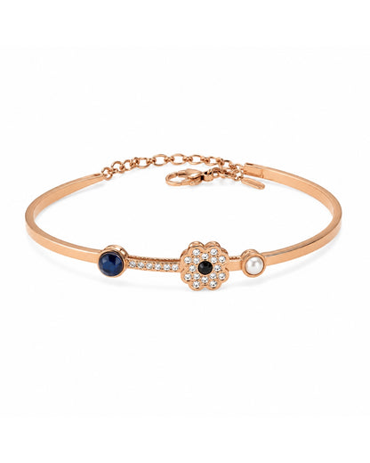 A rose gold bangle bracelet featuring a central flower motif with a black center, surrounded by clear crystals, flanked by a dark blue oval stone and a small white pearl, on a clean white background.