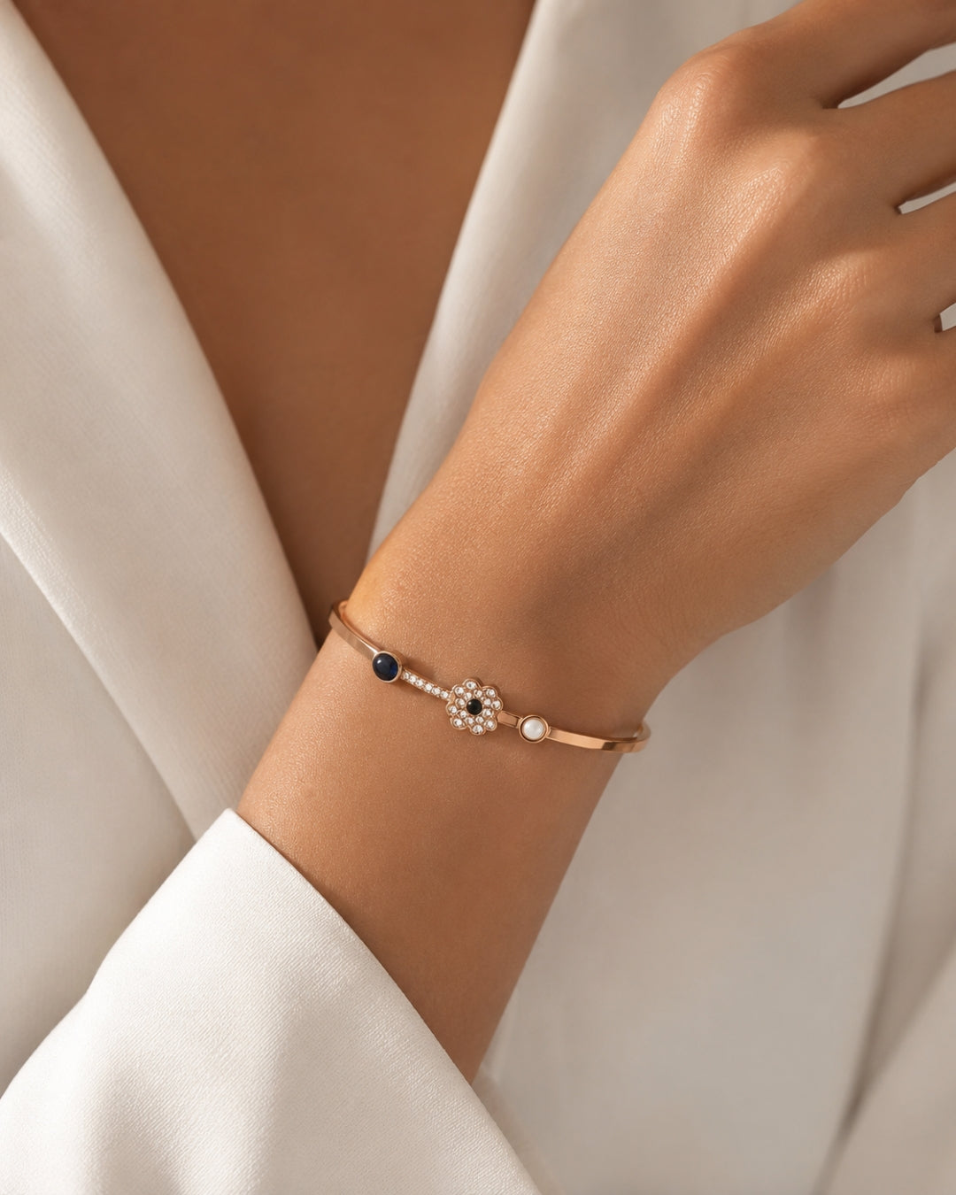 A delicate rose gold bangle bracelet with a central floral cluster of clear stones, a small white pearl, and a dark blue bead, shown worn on a woman's wrist.