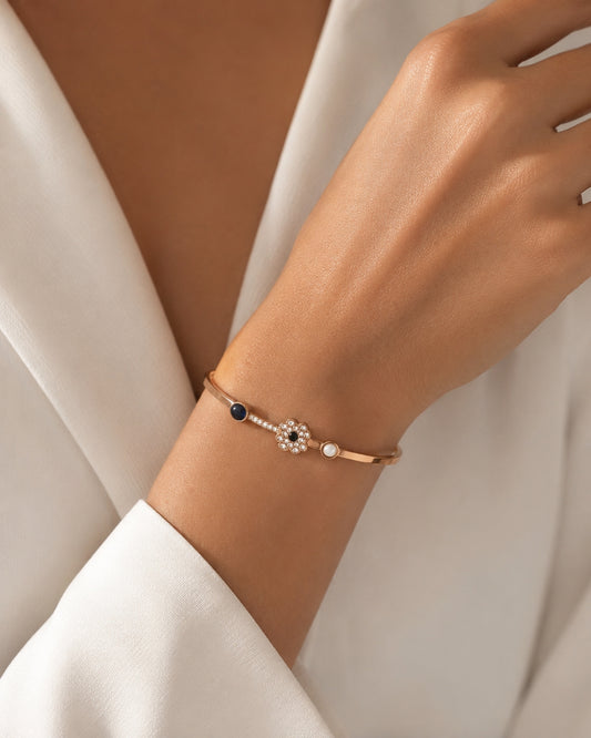 A delicate rose gold bangle bracelet with a central floral cluster of clear stones, a small white pearl, and a dark blue bead, shown worn on a woman's wrist.