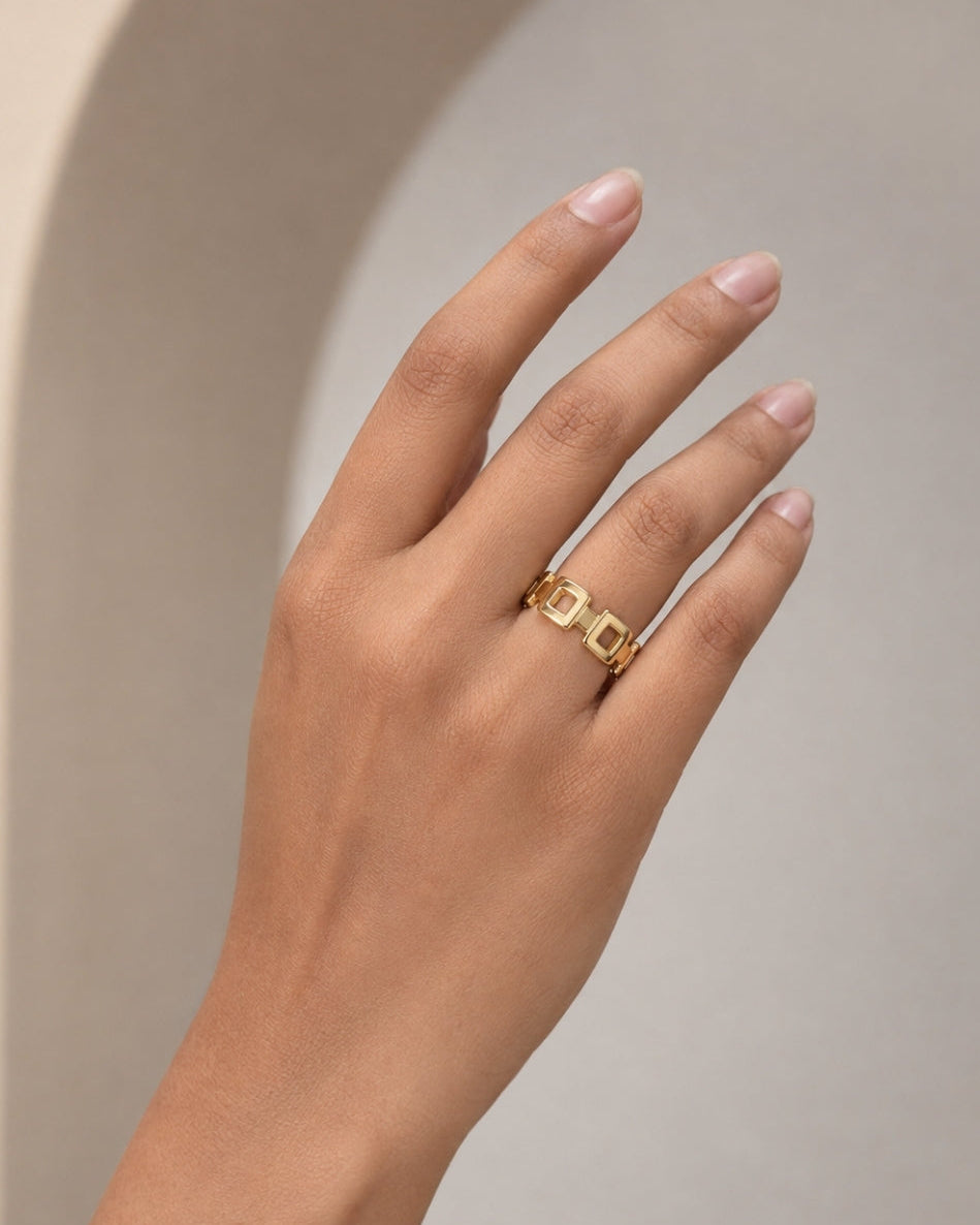 A close-up of a hand wearing a delicate gold ring with a repeating geometric pattern, set against a soft beige background with a subtle arch shadow.