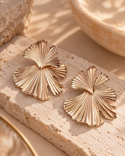 A pair of gold-toned fan-shaped earrings with a textured, ribbed surface, displayed on a beige travertine stone slab.