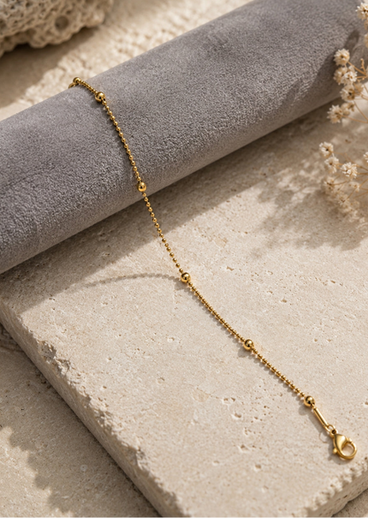 A delicate gold chain bracelet with small bead links and a lobster clasp, draped over a gray velvet cylindrical display roll on a textured beige stone surface with dried flowers.