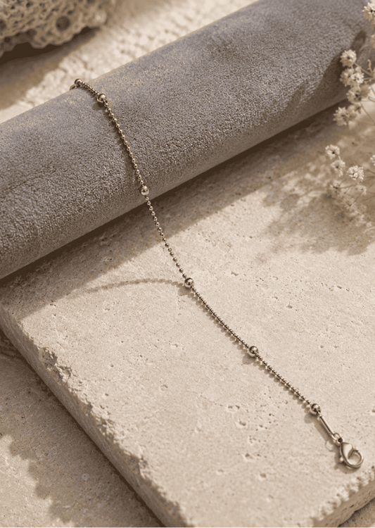 A delicate gold chain bracelet with small bead links and a lobster clasp, draped over a gray velvet cylindrical display roll on a textured beige stone surface with dried flowers.