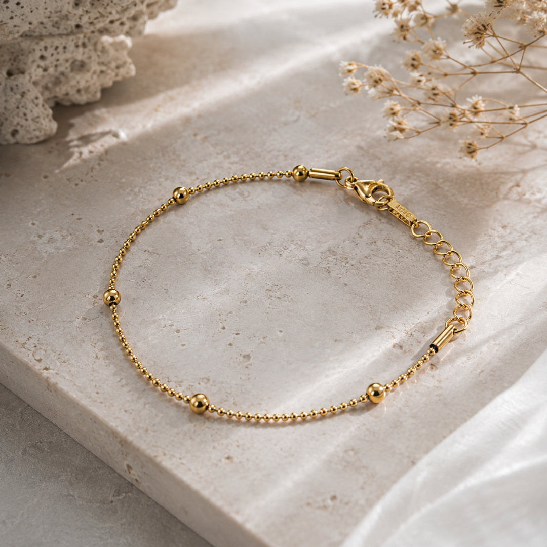 A delicate gold chain bracelet with small bead accents and a lobster clasp, displayed on a textured beige stone surface with dried flowers in the background.