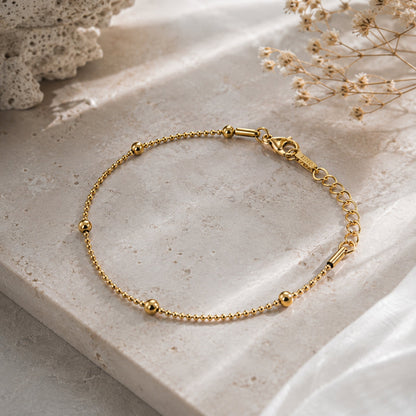 A delicate gold chain bracelet with small bead accents and a lobster clasp, displayed on a textured beige stone surface with dried flowers in the background.