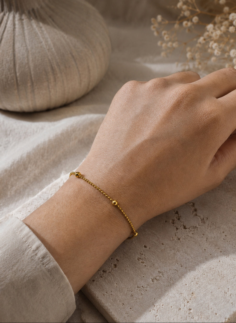 A delicate gold chain bracelet with small bead accents worn on a person's wrist, shown against a neutral beige fabric and stone background with dried flowers.