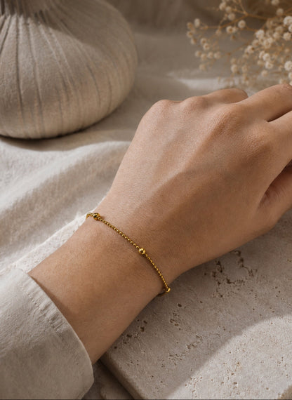 A delicate gold chain bracelet with small bead accents worn on a person's wrist, shown against a neutral beige fabric and stone background with dried flowers.