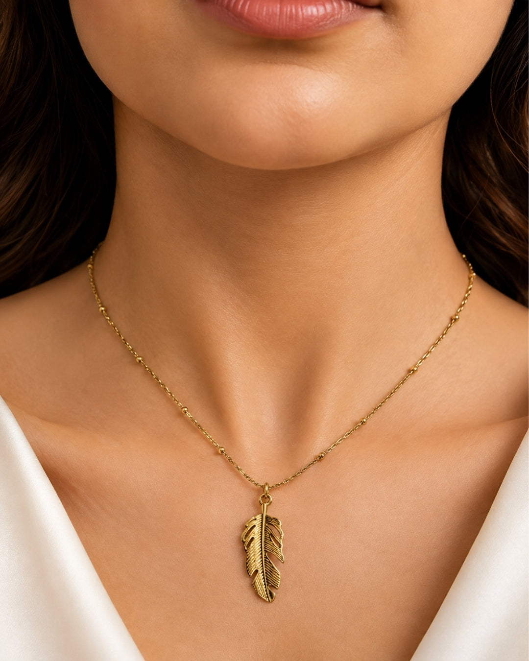 A close-up of a woman's neck and chest wearing a delicate gold chain necklace with a small feather pendant, styled with a white satin top.
