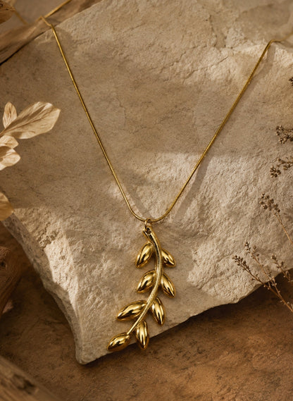 A gold leaf pendant necklace draped over a textured stone slab with dried botanical elements in the background.