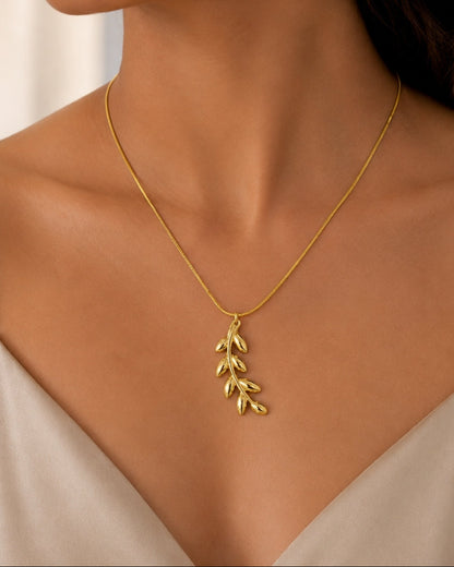 A close-up of a woman wearing a delicate gold necklace with a leafy branch pendant, styled with a beige camisole top.