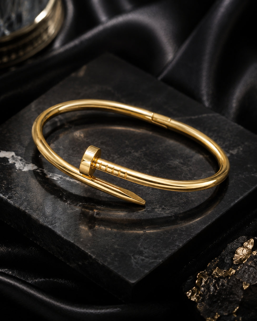 A polished gold-toned metal cuff bracelet with a distinctive screw-head closure, displayed on a dark textured stone slab with black satin fabric in the background.