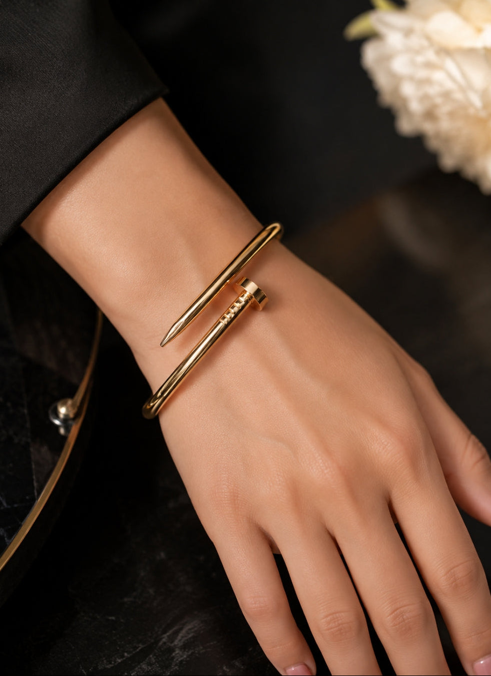 A close-up of a person's hand wearing a gold-toned wrap bangle bracelet with a textured cylindrical accent, set against a dark background with a blurred cream flower.