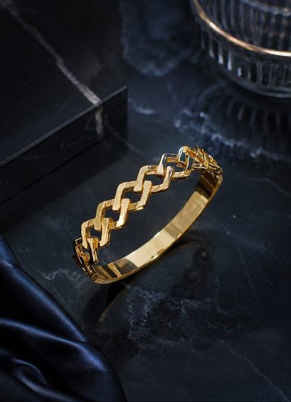A gold bangle bracelet with a repeating open geometric link pattern, displayed on a dark marble surface with a black box and dark fabric in the background.