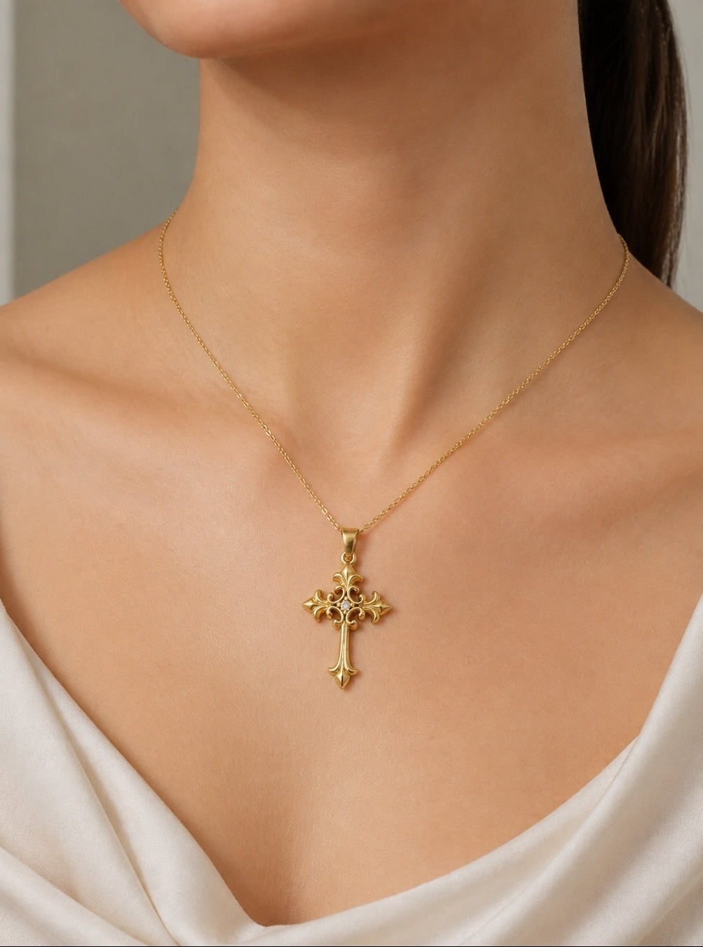 A close-up of a model wearing a delicate gold cross pendant necklace with a fine chain, styled with a white satin top.