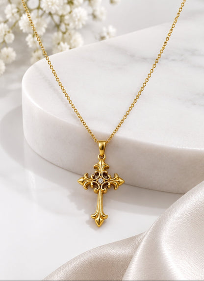 A gold cross pendant necklace with a delicate chain, displayed on a white marble pedestal with soft white flowers and satin fabric in the background.