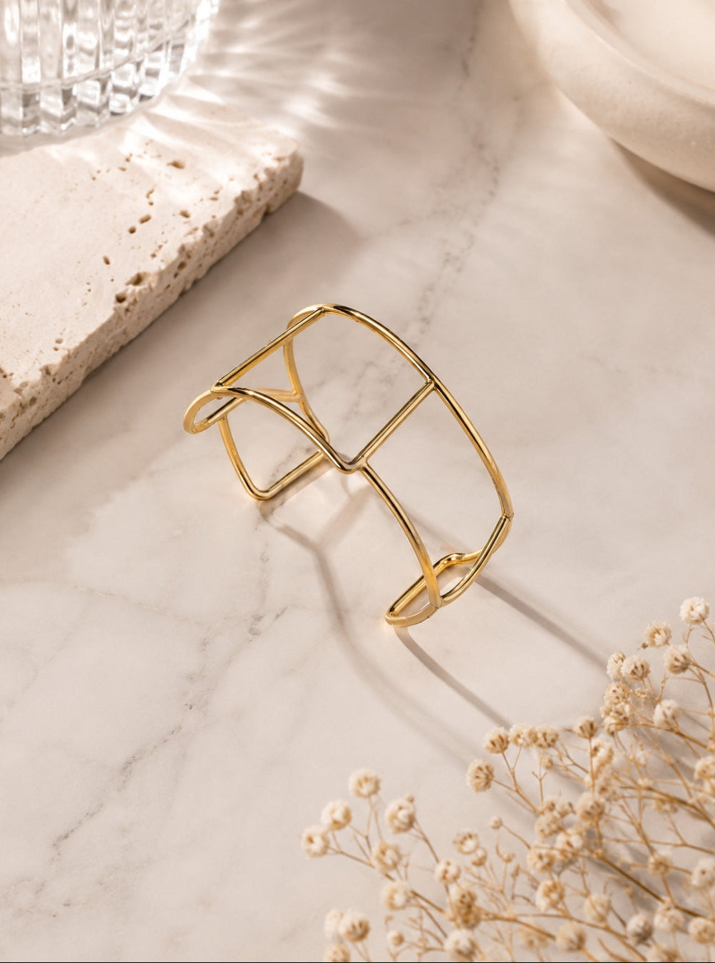A gold-toned open cuff bracelet with a geometric, cage-like design, displayed on a white marble surface with dried baby's breath flowers and neutral props.