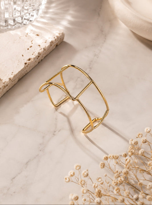 A gold-toned open cuff bracelet with a geometric, cage-like design, displayed on a white marble surface with dried baby's breath flowers and neutral props.