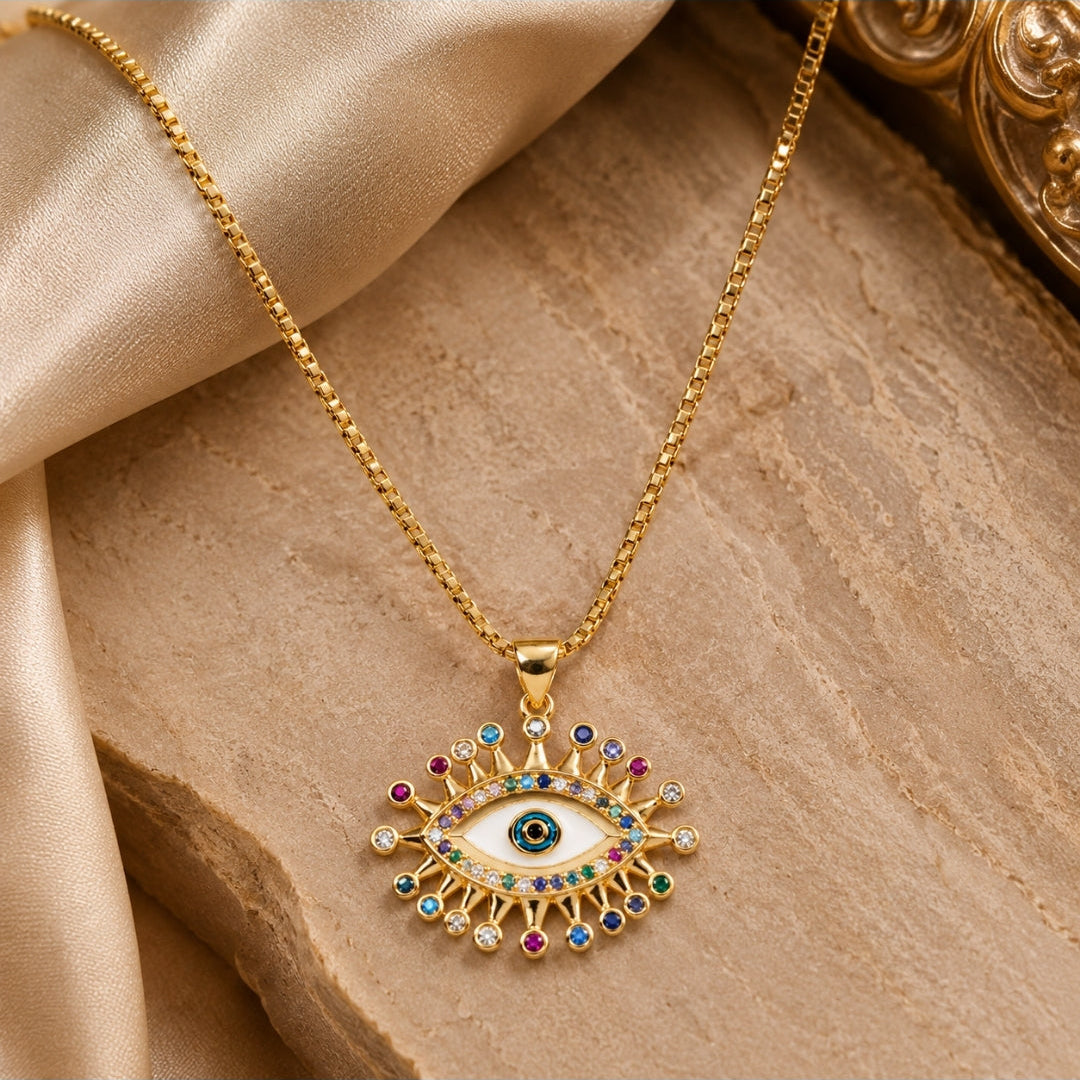 Gold-tone evil eye pendant necklace with a white enamel center and multicolored gem accents, displayed on draped beige satin fabric.