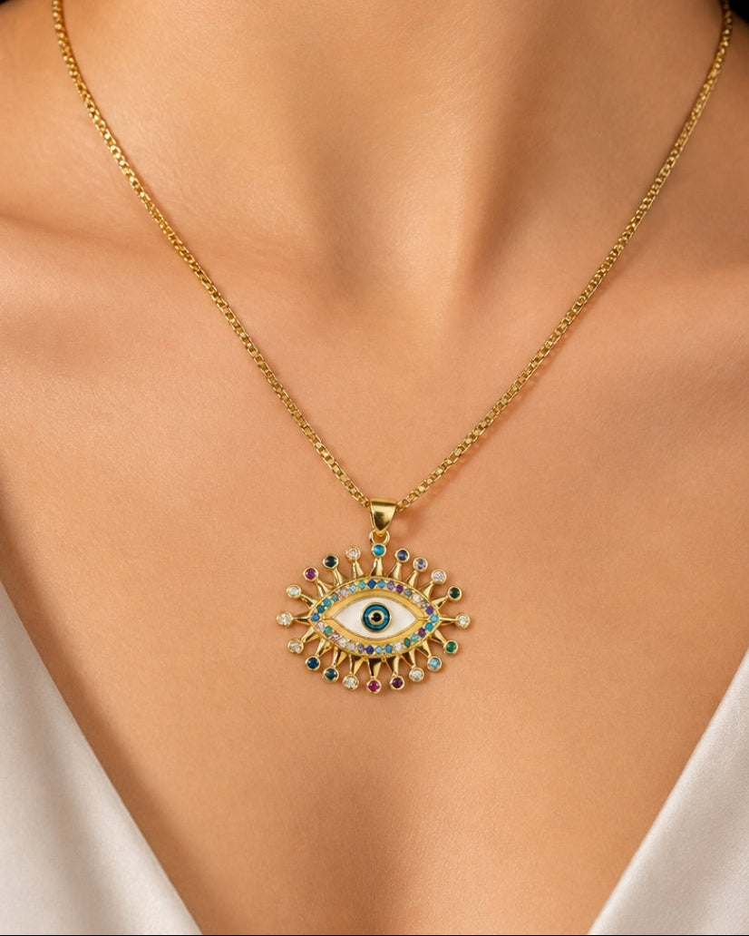 A gold chain necklace with an evil eye pendant, worn by a model. The pendant features a gold-tone frame with turquoise, red, and white enamel details.
