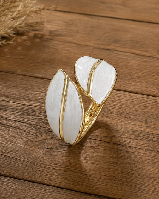 A gold-toned wrap-style ring with two large white enamel leaf-shaped inlays, photographed on a rustic wooden surface.
