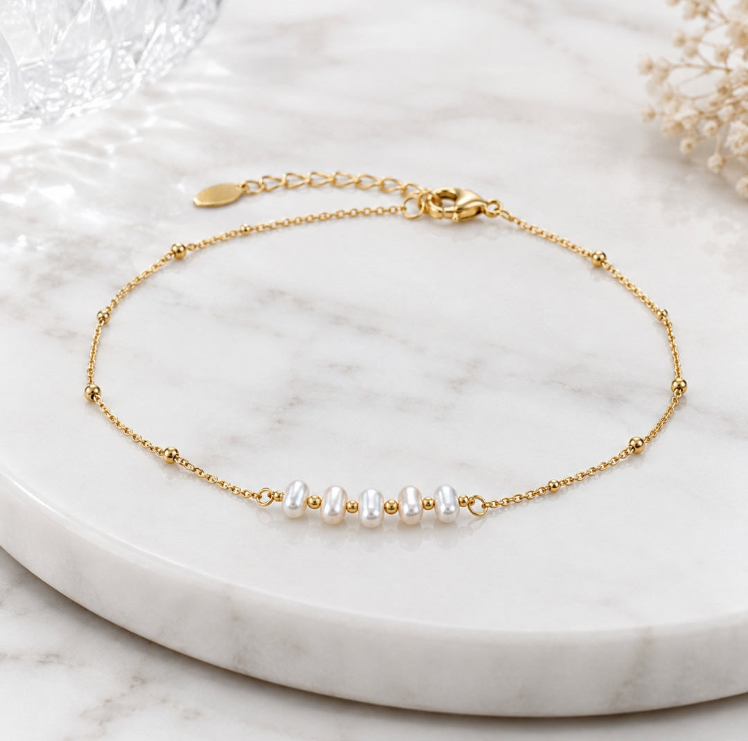 A delicate gold chain bracelet with five small white pearls and tiny gold beads, displayed on a white marble tray.