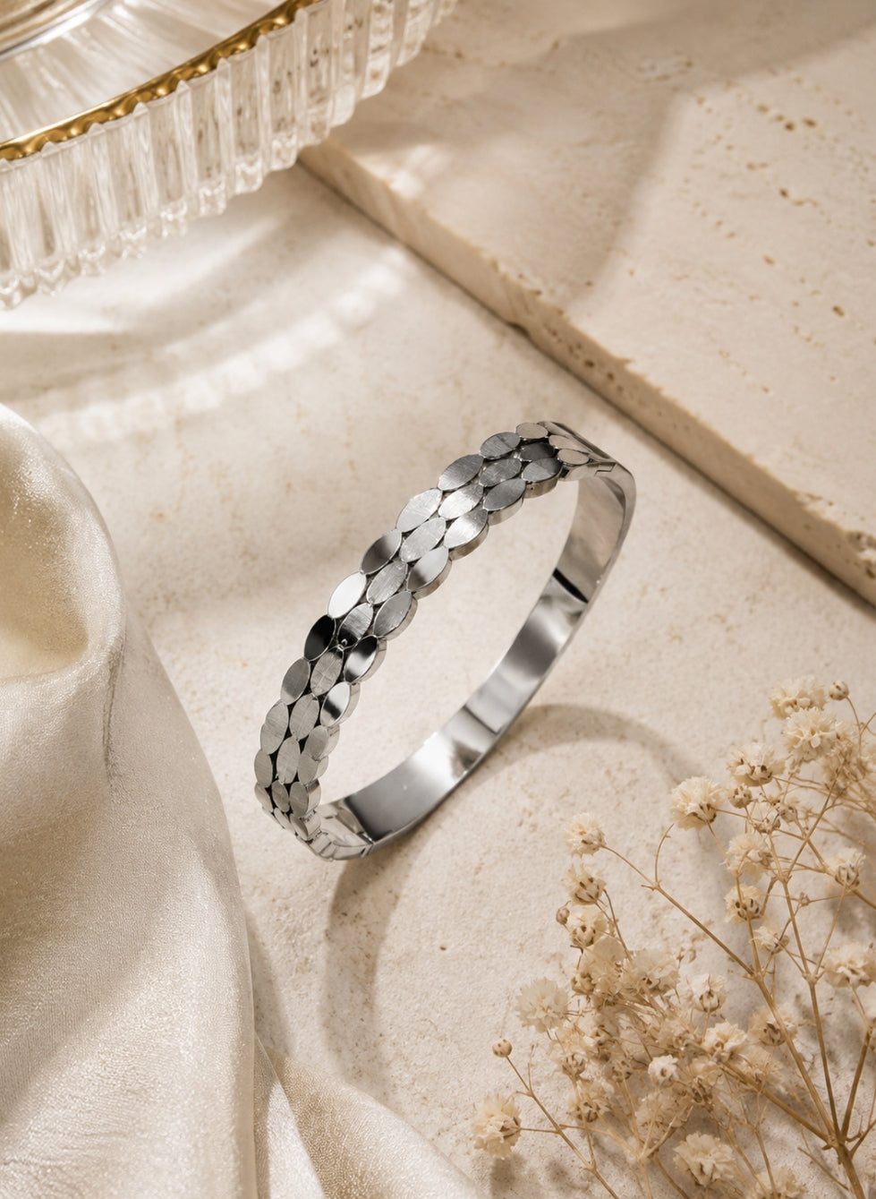 A polished silver bangle bracelet with a textured, overlapping petal-like pattern, displayed on a beige stone surface with draped cream fabric and dried baby's breath flowers.