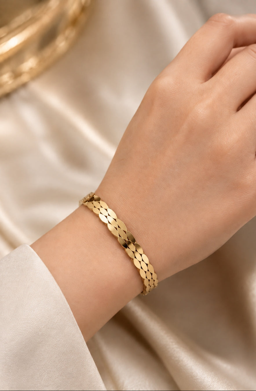 A close-up of a person's wrist wearing a gold chain-link bracelet with a textured, overlapping scale pattern, set against a soft beige satin fabric background.