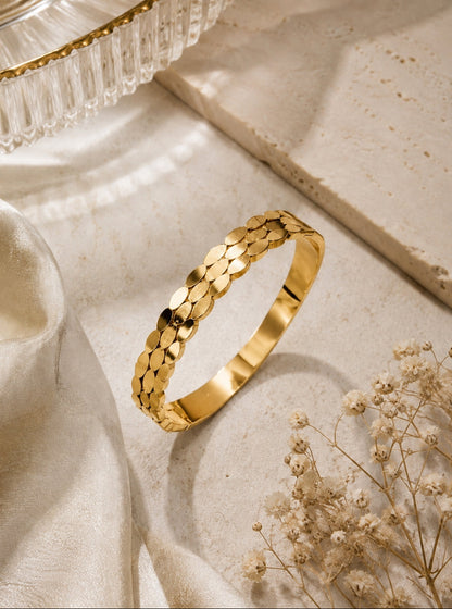 A gold-toned bangle bracelet with a textured, overlapping scale pattern, displayed on a light beige stone surface with draped cream fabric and dried flowers.