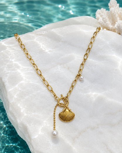A gold-tone chain necklace with a shell-shaped pendant, a small dangling bead chain, and pearl-like beads, displayed on a white stone with turquoise water and coral in the background.