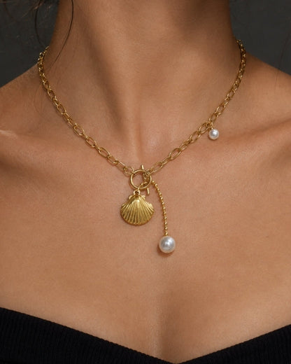 A close-up of a woman wearing a delicate gold chain necklace with a shell-shaped pendant and a small pearl drop, paired with a black off-the-shoulder top against a dark gray background.