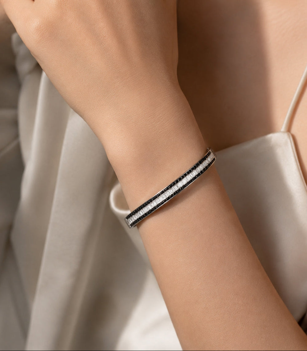 A close-up of a person's wrist wearing a slim black and white striped bangle bracelet, paired with a light-colored satin top.