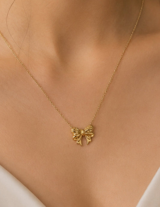 A delicate gold chain necklace with a small bow-shaped pendant, worn on a model's neck over a white top.