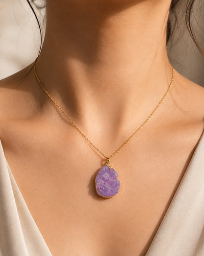 A gold chain necklace with a teardrop-shaped purple druzy stone pendant, worn by a model in a close-up shot.