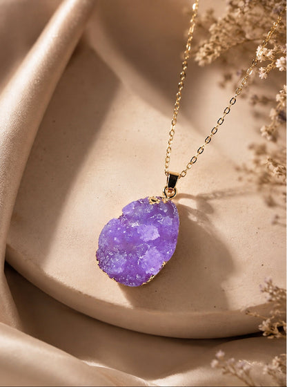 A gold chain necklace with a polished purple druzy stone pendant, styled on a beige ceramic dish with soft satin fabric and dried flowers.