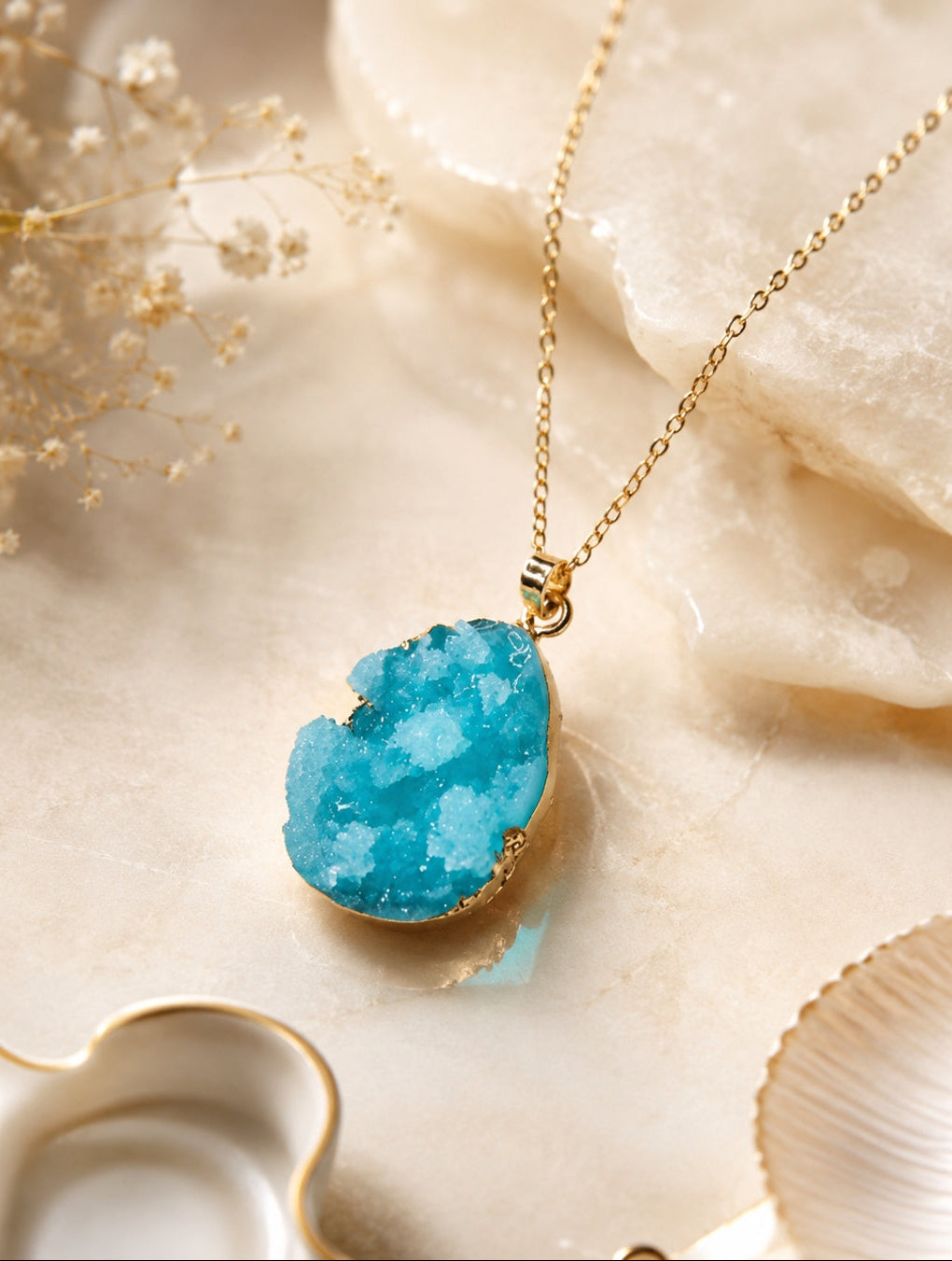 A gold chain necklace with a teardrop-shaped turquoise blue druzy stone pendant, styled on a beige marble surface with dried flowers and crystals.