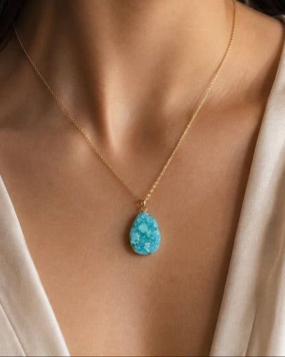 A close-up of a woman wearing a delicate gold chain necklace with a teardrop-shaped turquoise pendant, paired with a cream satin top.