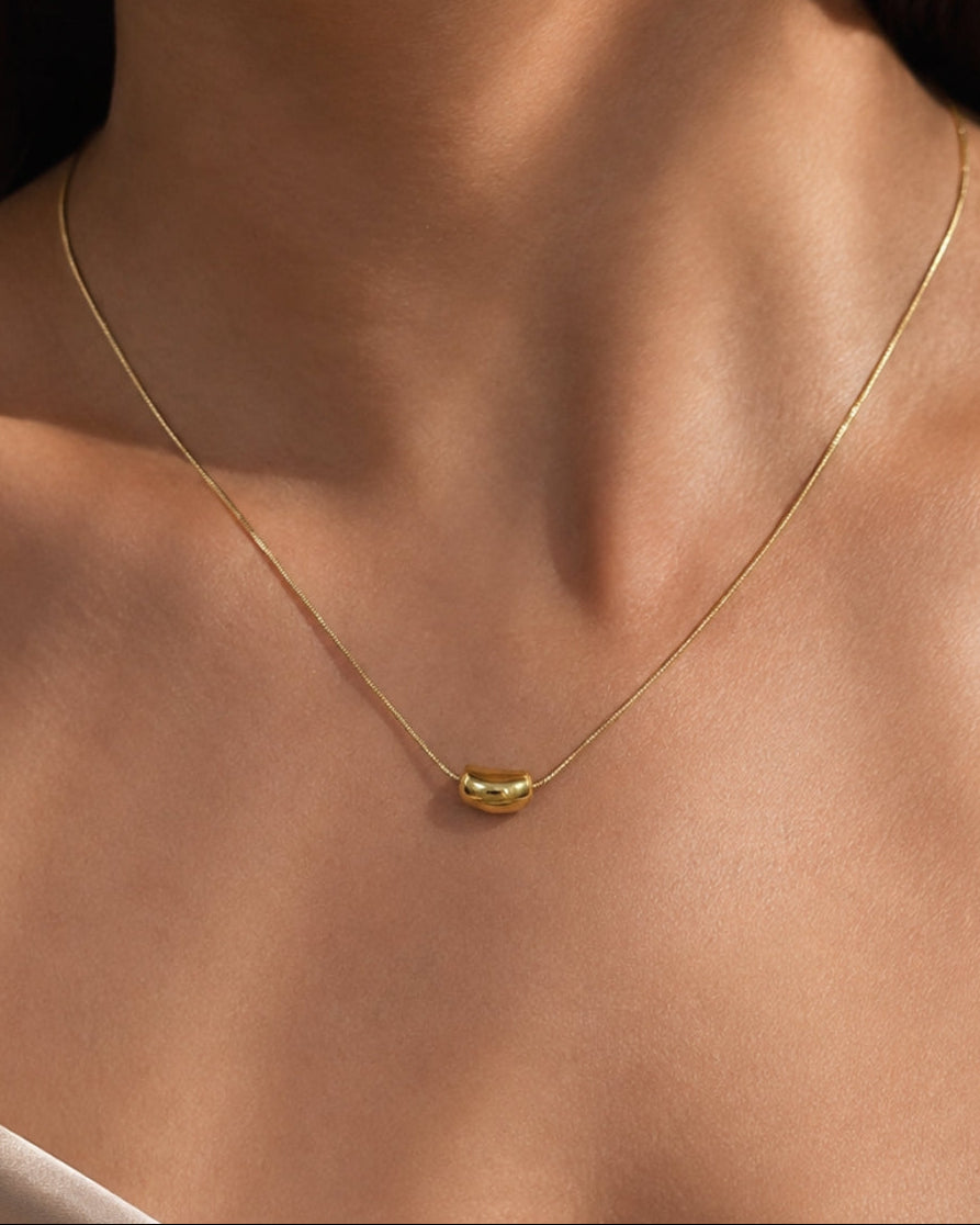 A close-up of a delicate gold chain necklace worn by a model, featuring a small polished gold capsule pendant at the center of the chest.