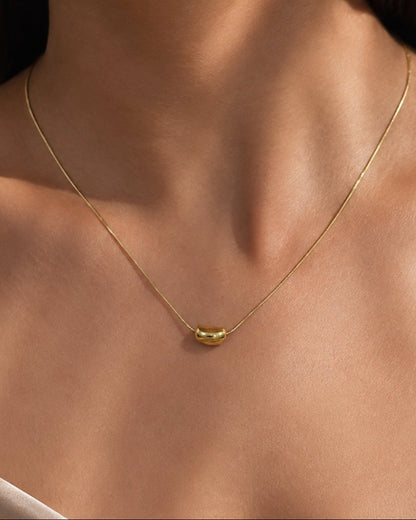 A close-up of a delicate gold chain necklace worn by a model, featuring a small polished gold capsule pendant at the center of the chest.
