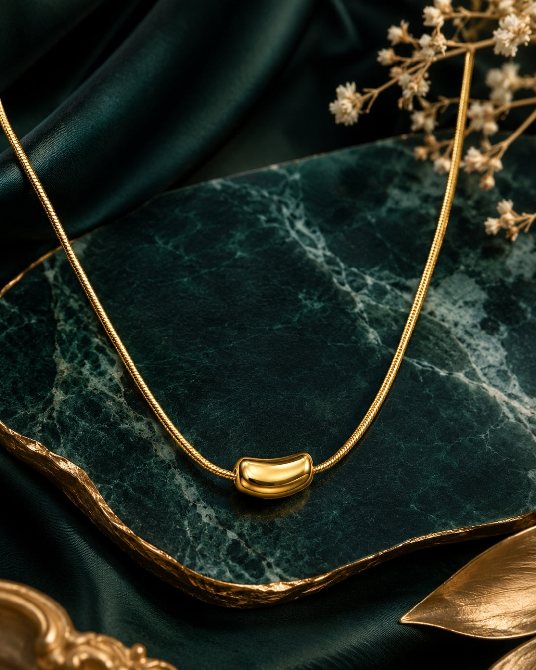 A gold snake chain necklace with a small curved, polished gold pendant, displayed on a dark green marble tray with dark green satin fabric and dried flowers.