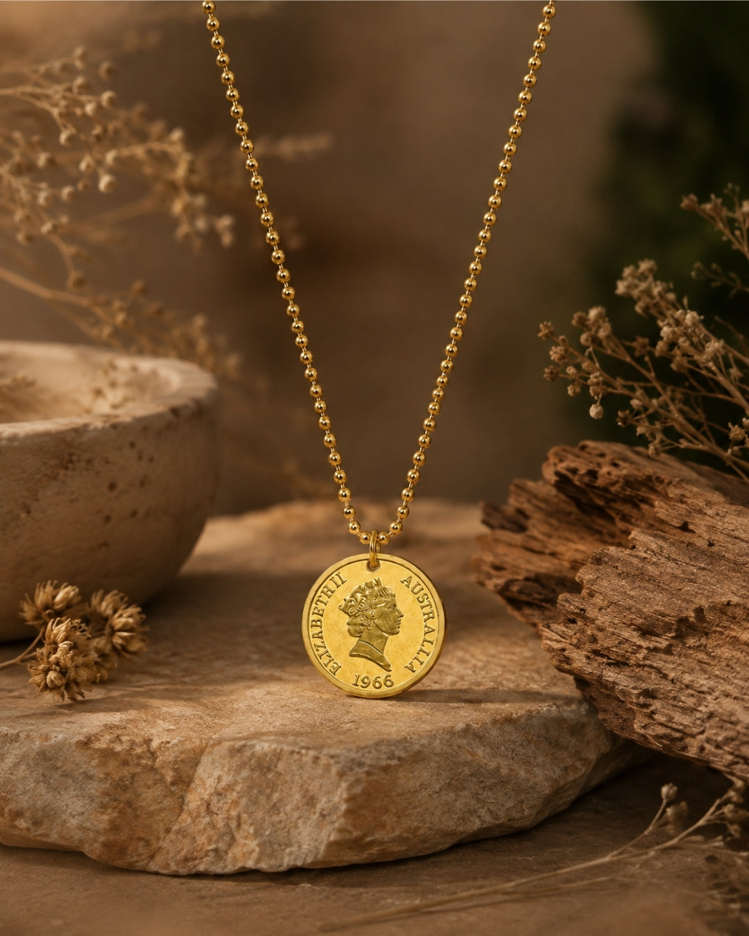 A gold coin pendant necklace displayed on a stone surface with dried flowers and rustic wood props. The pendant is a round gold coin featuring a profile portrait and the text 'ELIZABETH II DEI GRATIA 1966'.