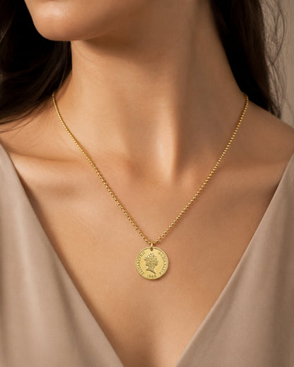 A close-up of a model wearing a delicate gold chain necklace with a round, coin-style pendant featuring an engraved profile.